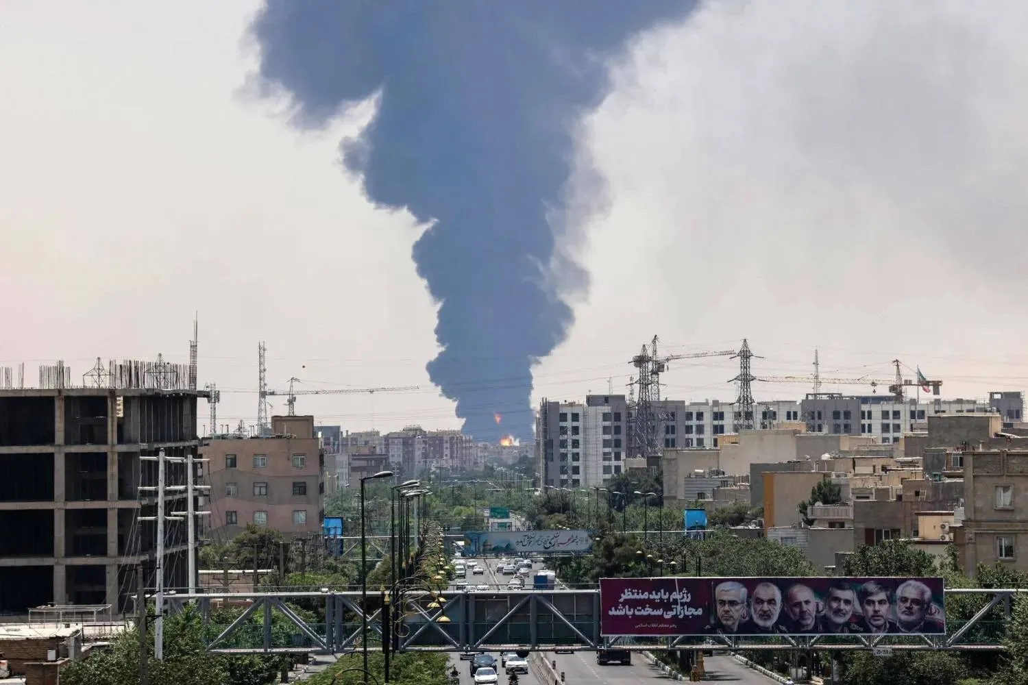 Portraits of Iranian military generals and nuclear scientists, killed in Israel's June 13 attack are displayed above a road, as a plume of heavy smoke and fire rise from an oil refinery in southern Tehran, after it was hit in an overnight Israeli strike, on June 15, 2025. (AFP) 