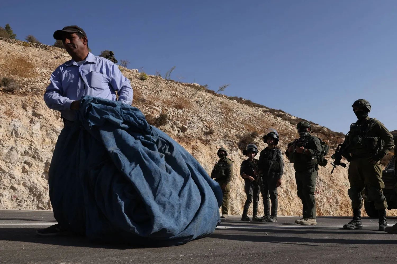فلسطيني يحمل كيساً من الزيتون وفي الخلفية قوات أمن إسرائيلية في الضفة الغربية (أ.ف.ب)