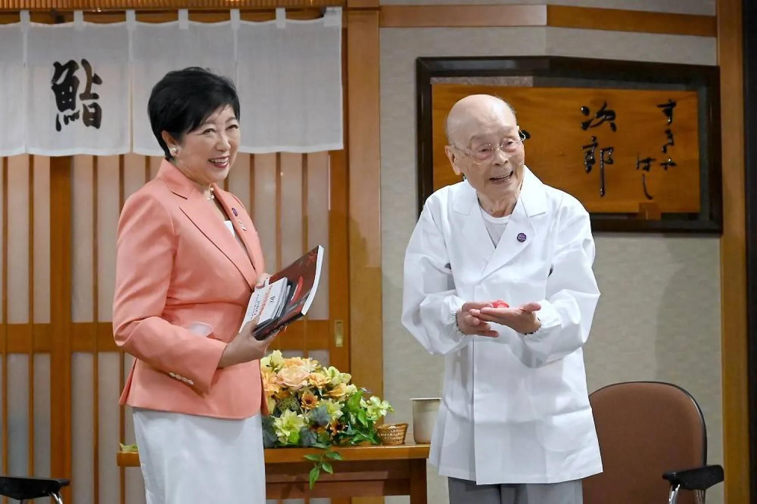 In this photo released by Bureau of Social Welfare, Tokyo Metropolitan Government, sushi legend Jiro Ono, right, shows a gift from Tokyo Gov. Yuriko Koike to celebrate his 100th birthday in front of his restaurant, Sukiyabashi Jiro in Tokyo on Sept. 18, 2025. (Bureau of Social Welfare, Tokyo Metropolitan Government via AP)