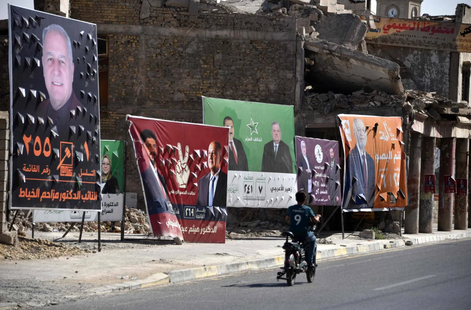 رجل يقود دراجته الهوائية أمام ملصقات تُظهر مرشحين للانتخابات العراقية بمدينة الموصل القديمة (أ.ف.ب)
