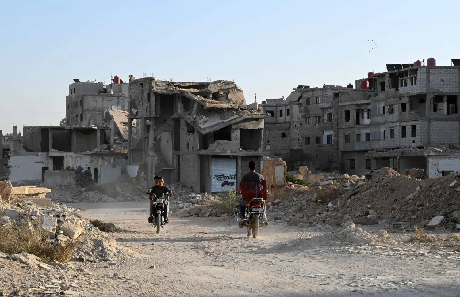Şam'ın dış mahallelerinden Derayya'da yıkılmış binaların önünde motosikletleriyle geçen insanlar, 28 Ekim 2025 (AFP)