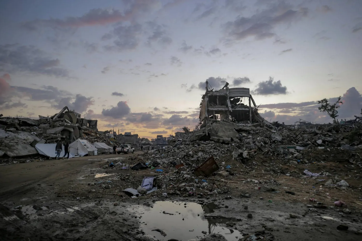 فلسطينيون يسيرون بالقرب من مبانٍ مدمرة في يوم ممطر شرق مدينة غزة - 16 نوفمبر 2025 (إ.ب.أ)