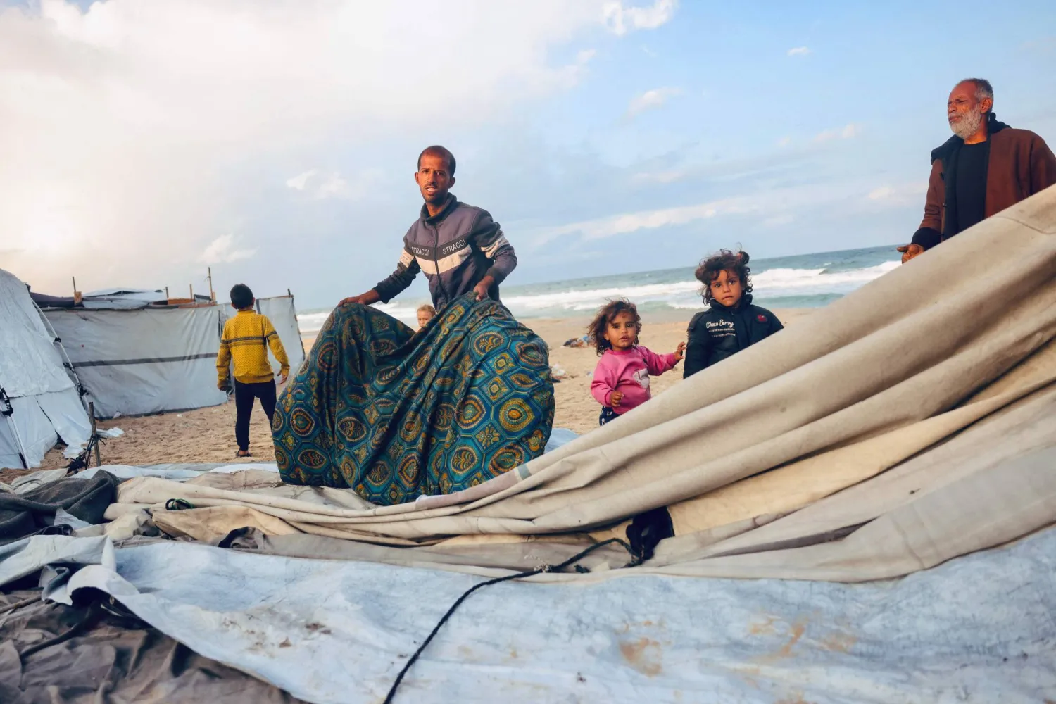 رجل فلسطيني وأطفاله يُعدّلون قماش مأواهم من المطر في مخيم مؤقت للنازحين الفلسطينيين بدير البلح وسط قطاع غزة (أ.ف.ب)