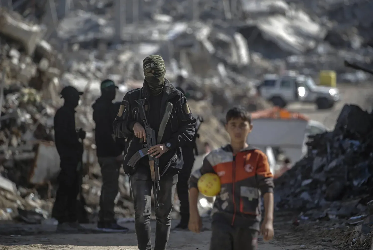 Gazze Şeridi'nin kuzeyindeki Cibaliye Mülteci Kampı’nda İsrailli rehinelerin cesetlerini arayan İzzeddin el-Kassam Tugayları savaşçılarının yanında duran Filistinli bir çocuk, 1 Aralık 2025 (EPA)