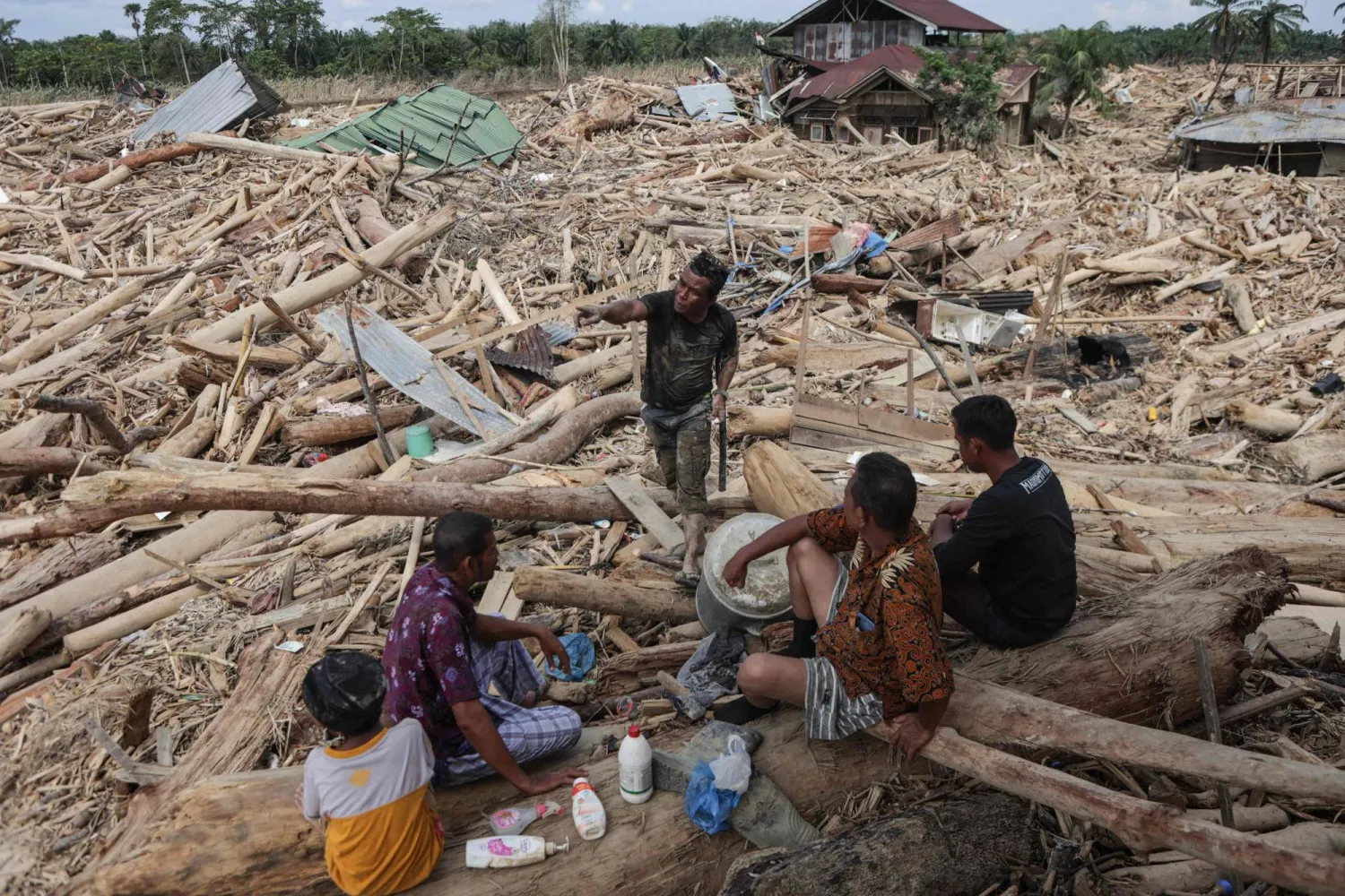 Residents rest as they search for the remains of their house, buried under piles of uprooted trees swept by the flash flood, in Lintang Baru village in Aceh Tamiang, northern Sumatra, on December 11, 2025. (Photo by Aditya Aji / AFP)