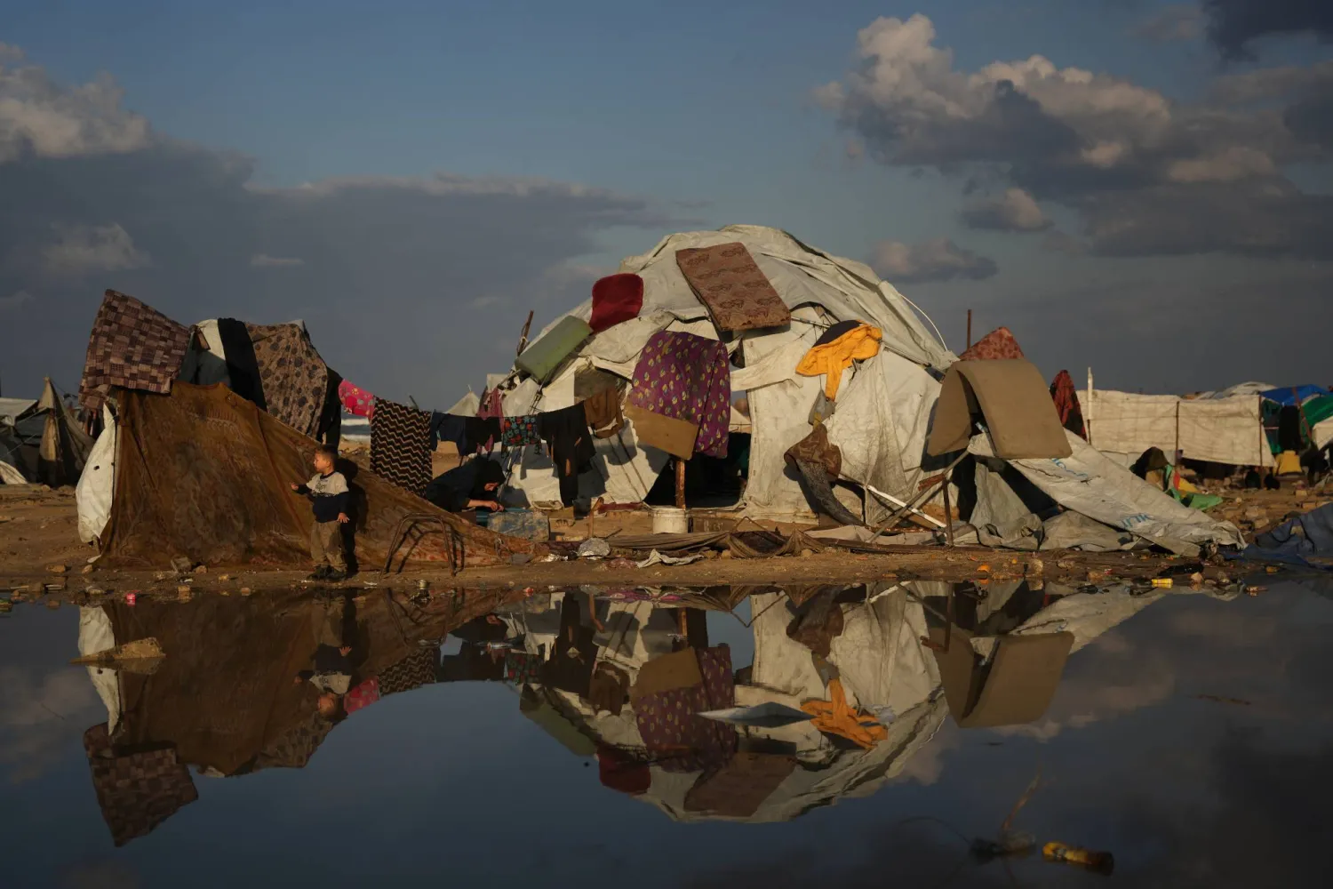 طفل فلسطيني يقف بجانب والدته وهي تغسل الملابس بينما تُعلق المراتب لتجف فوق خيمتهم في مخيم مؤقت للنازحين أقيم على شاطئ مدينة غزة (أ.ب)