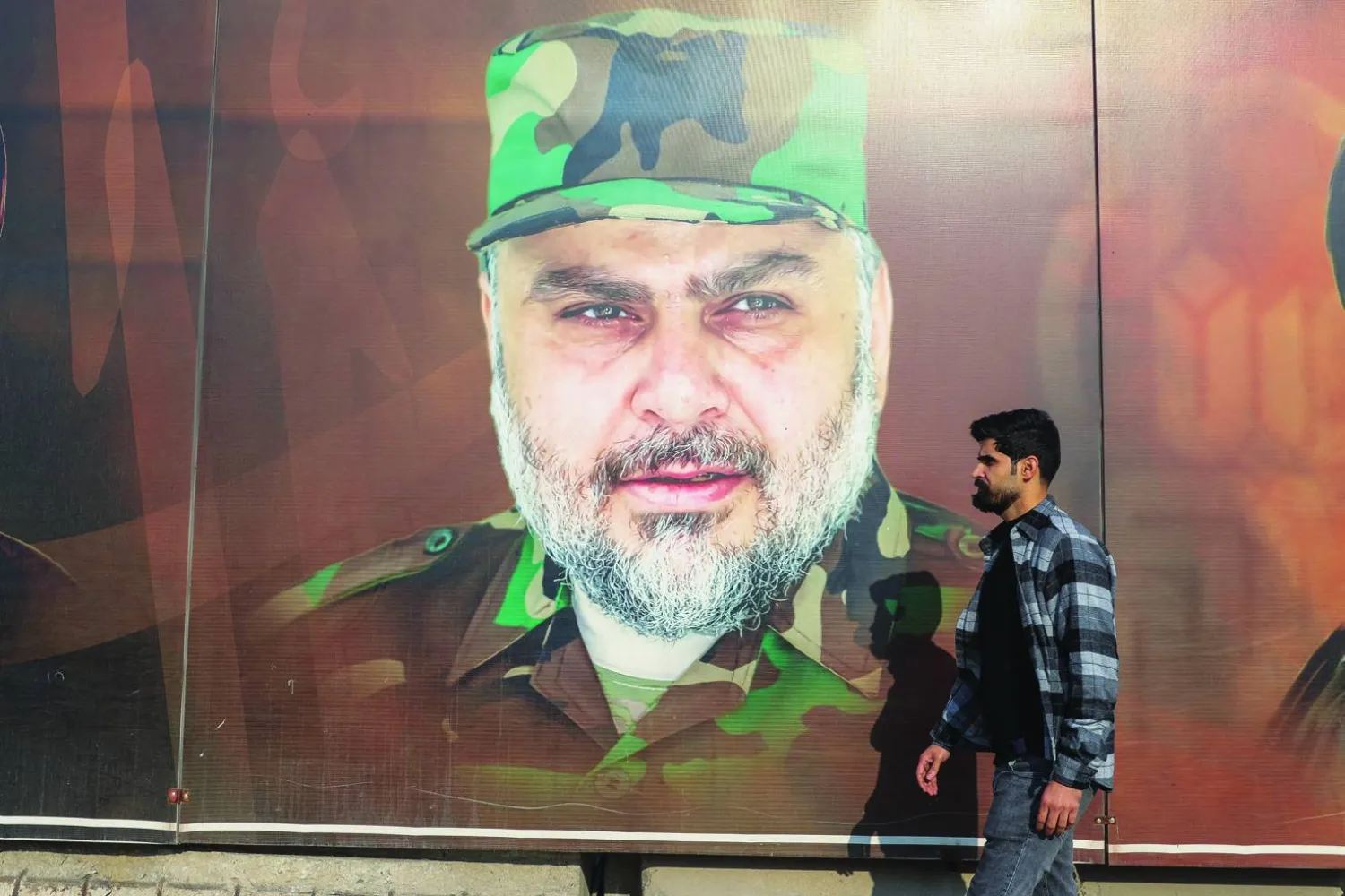 عراقي بأحد شوارع بغداد يمر أمام صورة لمقتدى الصدر مرتدياً الزي العسكري الخاص بـ«سرايا السلام»  (أ.ف.ب)