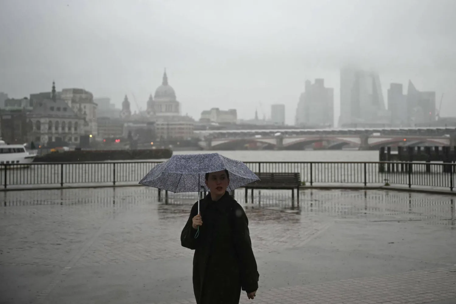 A pedestrian shelters from the rain while walking along the South Bank with the River Thames and the office buildings of the City Of London shrouded in fog on December 18, 2025. (Photo by JUSTIN TALLIS / AFP)