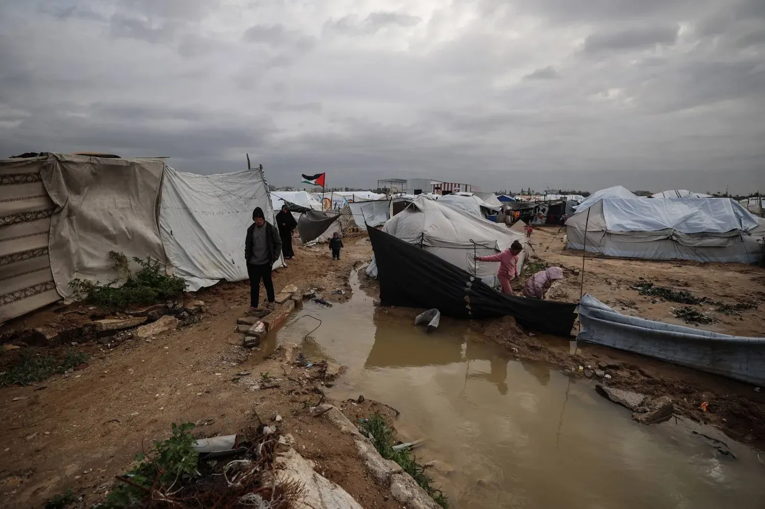 Gazze Şeridi'nin orta kesimlerindeki el-Bureyc Mülteci Kampı’ndaki geçici barınakların arasında, yerinden edilmiş Filistinliler bir yağmur suyu birikintisinin yanında duruyorlar (AFP)