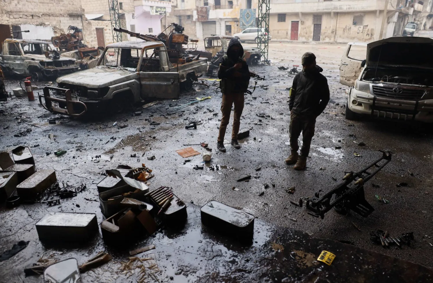 عنصران من قوات الأمن السورية يقفان وسط المركبات المحترقة والذخيرة المتناثرة على الأرص في حي الشيخ مقصود في حلب (أ.ب)