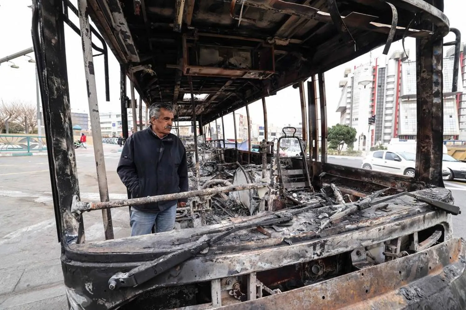 A man stands by the wreckage of a burnt bus bearing a banner (unseen) that reads "This was one of Tehran’s new buses that was paid for with the money of the people’s taxes,” in Tehran's Sadeghieh Square on January 15, 2026. (AFP)