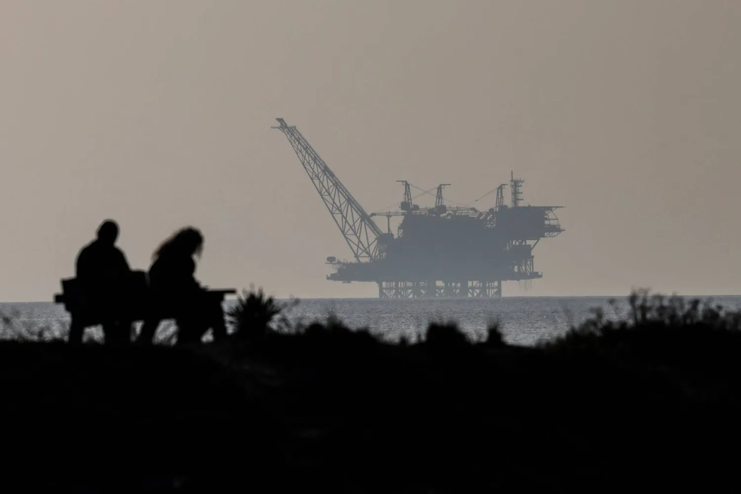 شخصان يجلسان على مقعد وخلفهم حقل غاز ليفياثان في محمية شاطئ هابونيم الطبيعية شمال إسرائيل (إ.ب.أ)