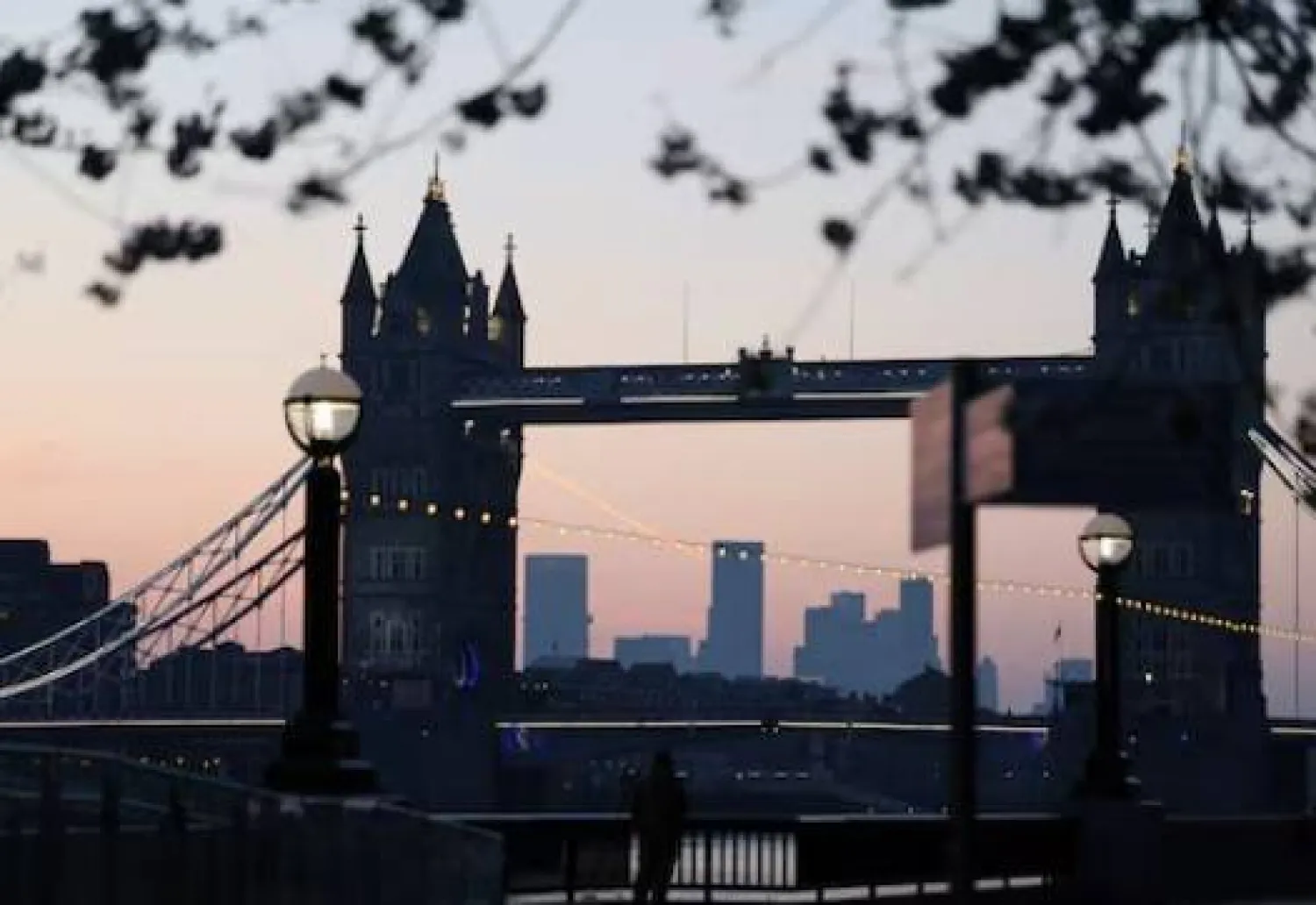 رجل يلتقط صوراً أثناء شروق الشمس خلف جسر البرج ومنطقة كاناري وارف المالية في لندن (رويترز)