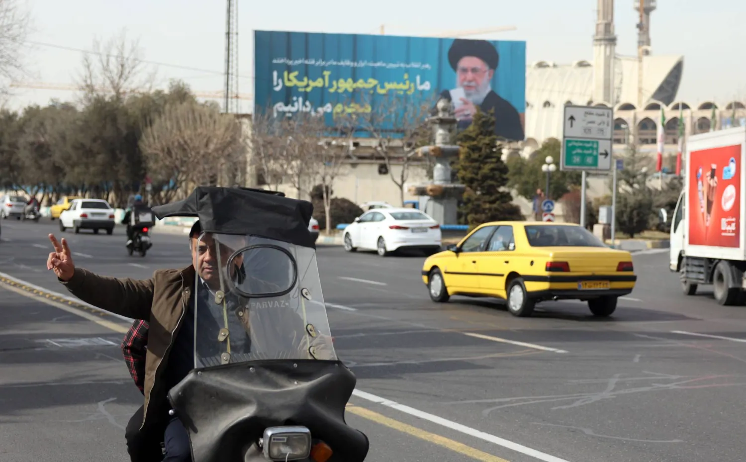 رجل إيراني يرفع علامة النصر أثناء مروره بجوار صورة للمرشد علي خامنئي في أحد شوارع طهران (إ.ب.أ)
