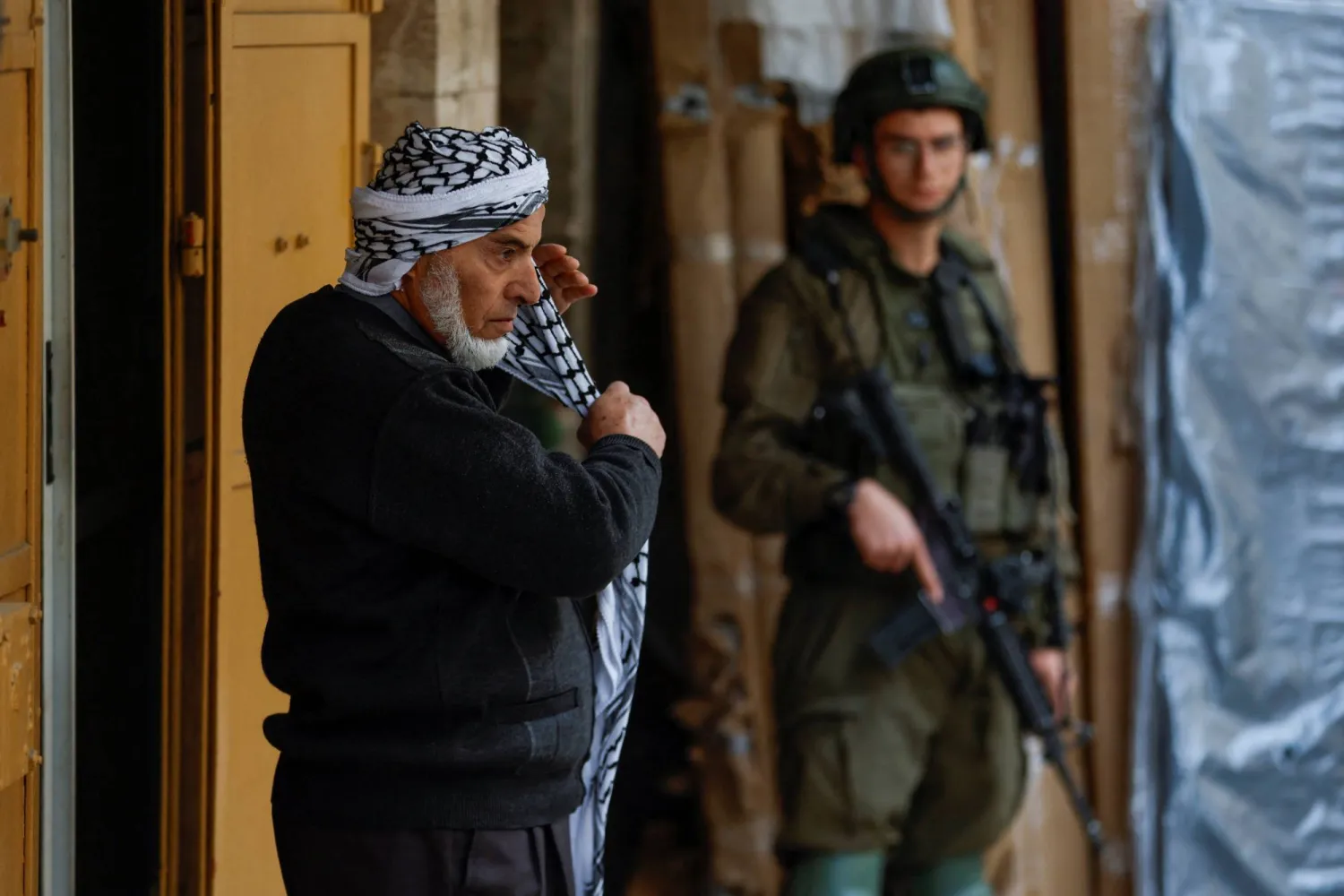 رجل فلسطيني يقف أمام جندي إسرائيلي في مدينة الخليل بالضفة الغربية المحتلة (رويترز)