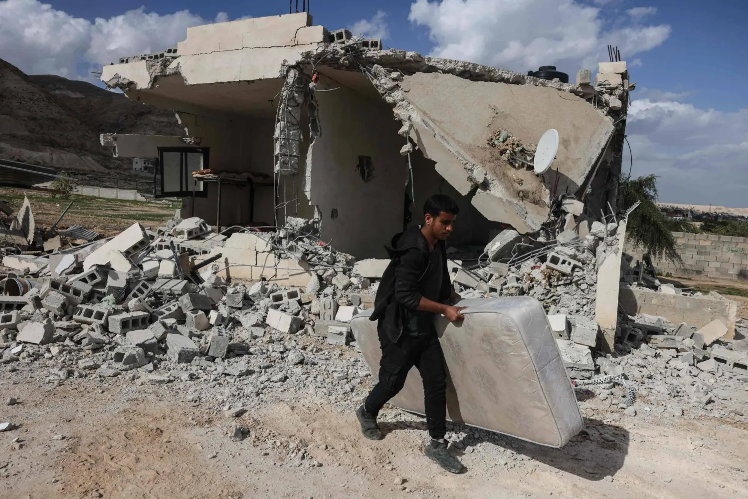 فلسطيني يحمل فراشاً ماراً بأنقاض منزل أفادت التقارير بهدمه على يد مستوطنين إسرائيليين في اليوم السابق بقرية على مشارف أريحا بالضفة الغربية المحتلة (أ.ف.ب)