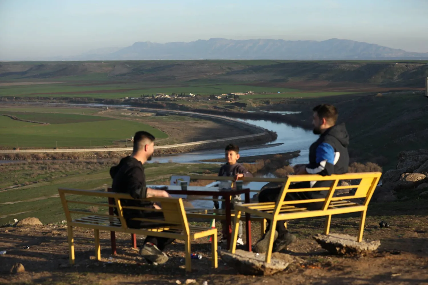 People sit outdoors surrounded by nature, with the Tigris river flowing in the background, following a long atmospheric depression, near the Syrian-Turkish border in Derik, Syria, February 16, 2026 REUTERS/Orhan Qereman

