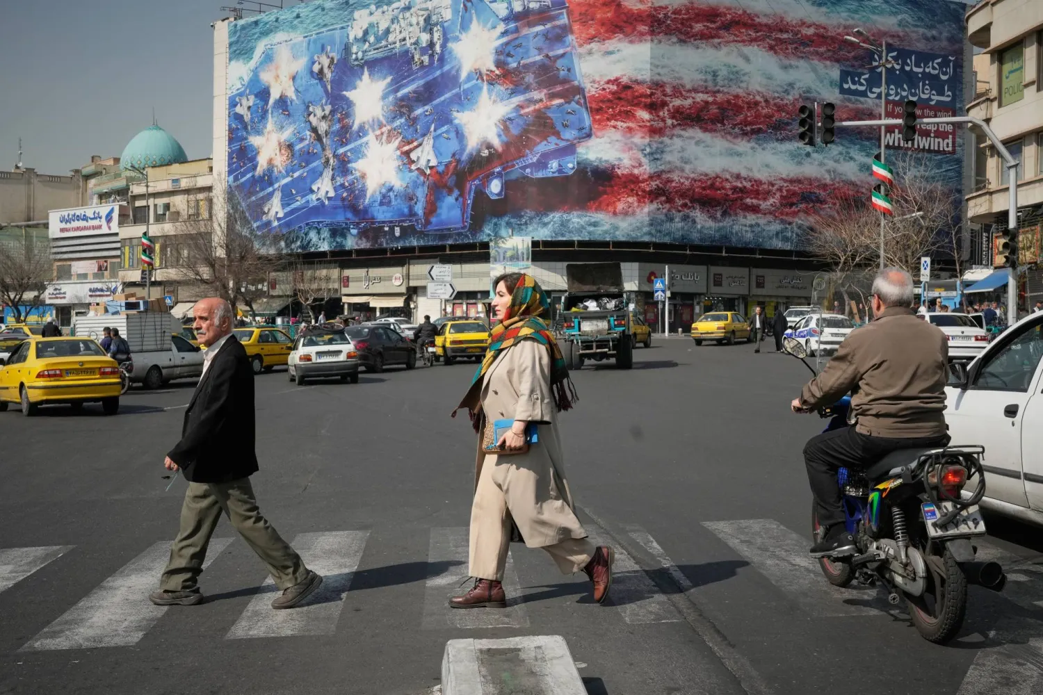 Pedestrians walk past a billboard depicting a US aircraft carrier with damaged fighter jets on its deck and a sign in Farsi and English reading, "If you sow the wind, you'll reap the whirlwind," in Tehran, Iran, Sunday, Feb. 22, 2026. (AP Photo/Vahid Salemi)