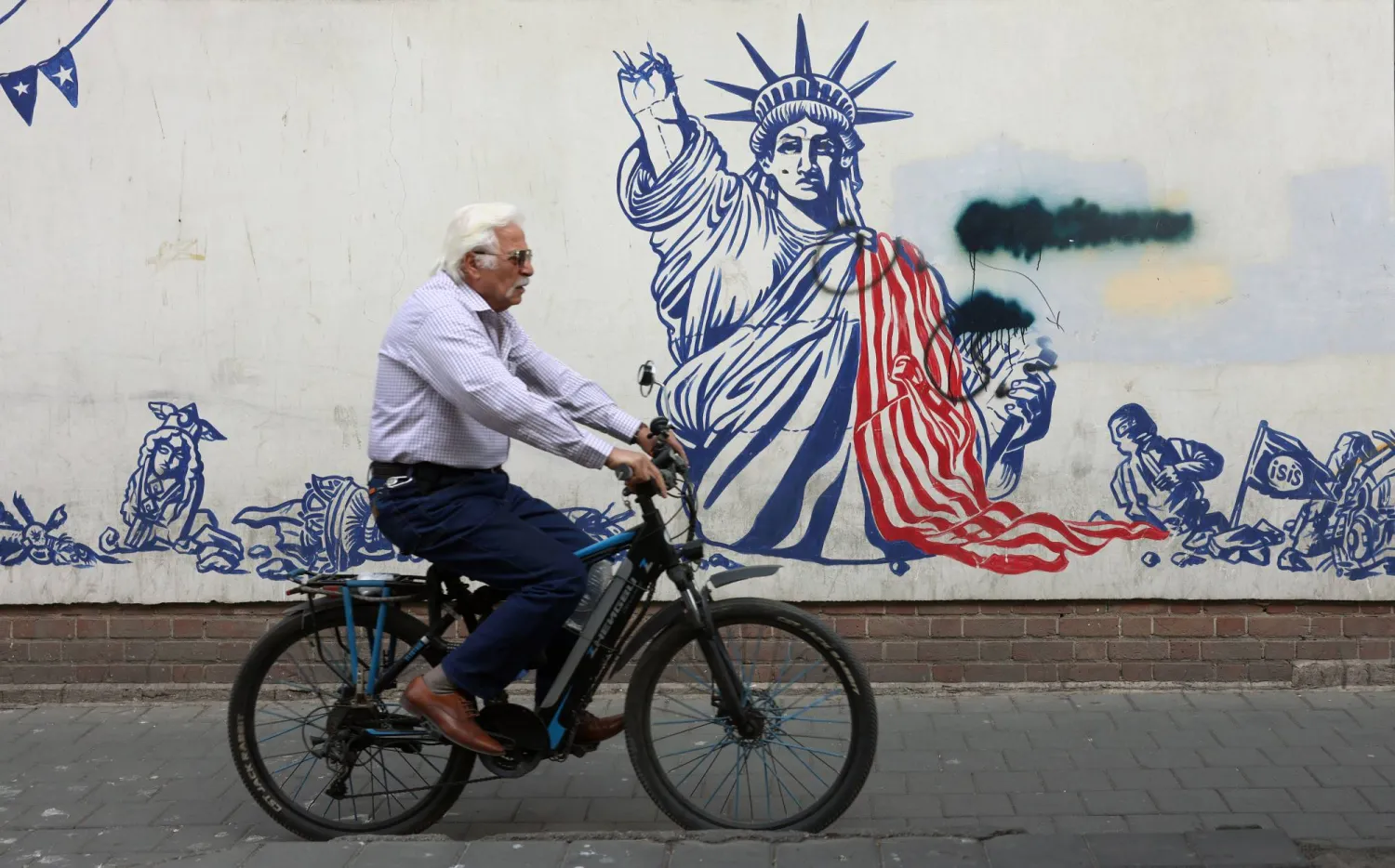 An elderly Iranian man rides a bicycle next to an anti-US mural in Tehran, Iran, 23 February 2026. EPA/ABEDIN TAHERKENAREH