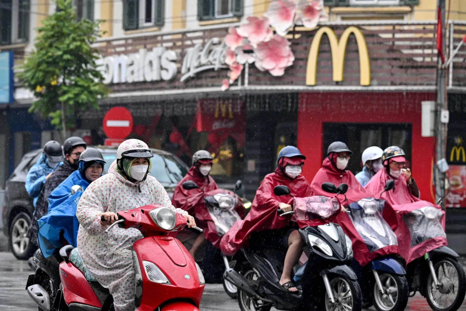 People wearing raincoats ride motorbikes through the rain past a fast-food restaurant in Hanoi on February 26, 2026. (Photo by Nhac NGUYEN / AFP)