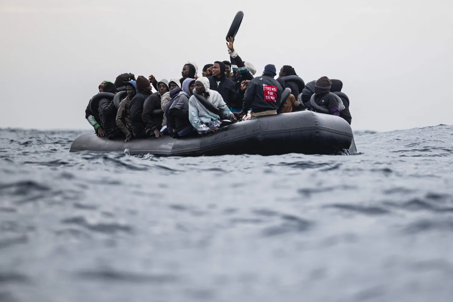(FILES) Migrants onboard a rubber boat wave and gesture as they wait to be rescued by crew members of the “Ocean Viking” rescue ship in the search-and-rescue zone in the Mediterranean Sea near the Libyan coast, on January 16, 2026. (Photo by Sameer Al-DOUMY / AFP)