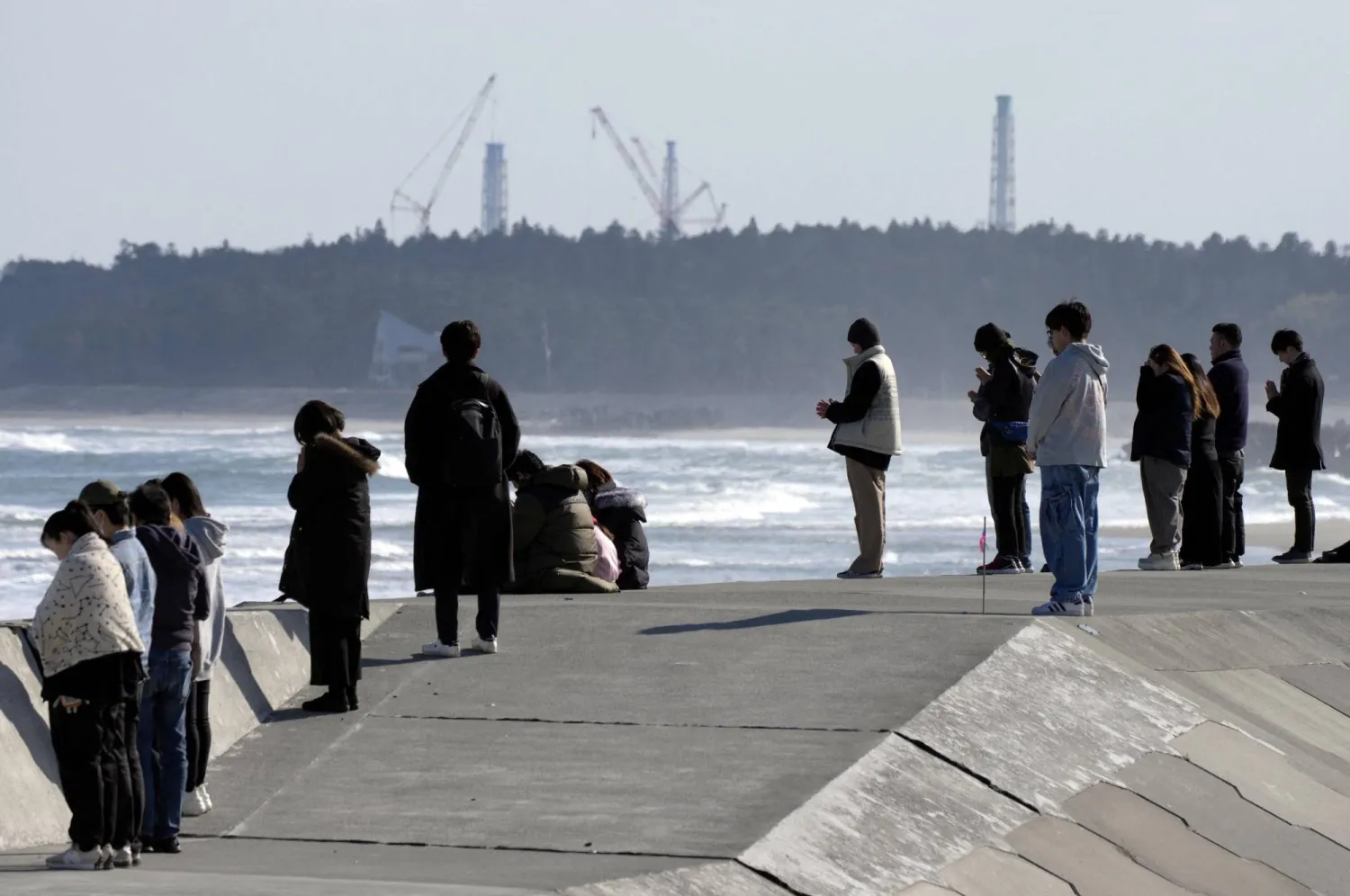 People observe a moment of silence towards the sea at 2:46 p.m. (05:46 GMT), the time when the 9.0-magnitude earthquake struck off Japan's coast in 2011, with the tsunami-crippled Fukushima Daiichi nuclear power plant seen in the background in Namie, Fukushima prefecture, Japan, March 11, 2026, to mark the 15-year anniversary of the 2011 earthquake and tsunami that killed thousands and set off a nuclear crisis. (Kyodo/via Reuters)