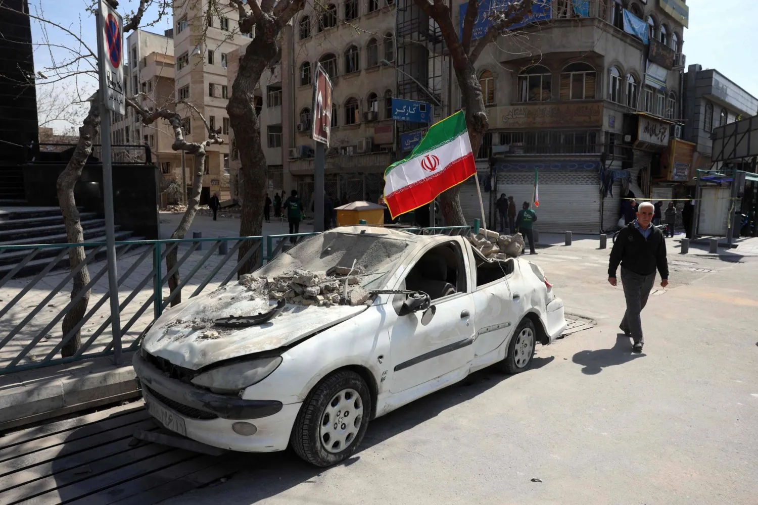 A man walks past an Iranian flag fluttering above the wreckage of a car in central Tehran, on March 4, 2026. (Photo by AFP)