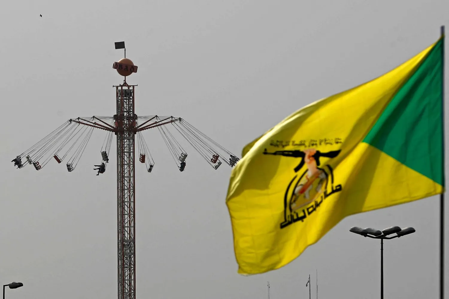 A flag of Iraq's pro-Iran paramilitary group Hezbollah Brigades (Kataib Hezbollah), flutters against the backdrop of people taking a swing ride at an amusement park during Eid al-Fitr celebrations, marking the end of the Muslim holy fasting month of Ramadan, in Baghdad on March 21, 2026. (Photo by AHMAD AL-RUBAYE / AFP)