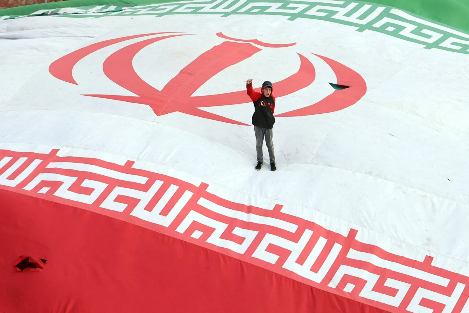 طفل يرفع قبضته بينما يقف على علم إيراني ضخم خلال جنازة (أ.ف.ب)