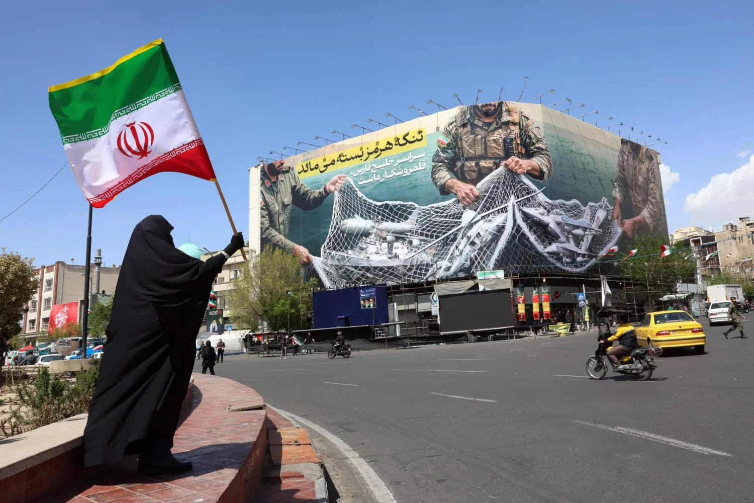A woman holds Iran’s national flag while standing near a billboard with a sentence reading ‘The Strait of Hormuz remains closed’ at the Enqelab Square in Tehran, on April 5, 2026. (Photo by AFP) 