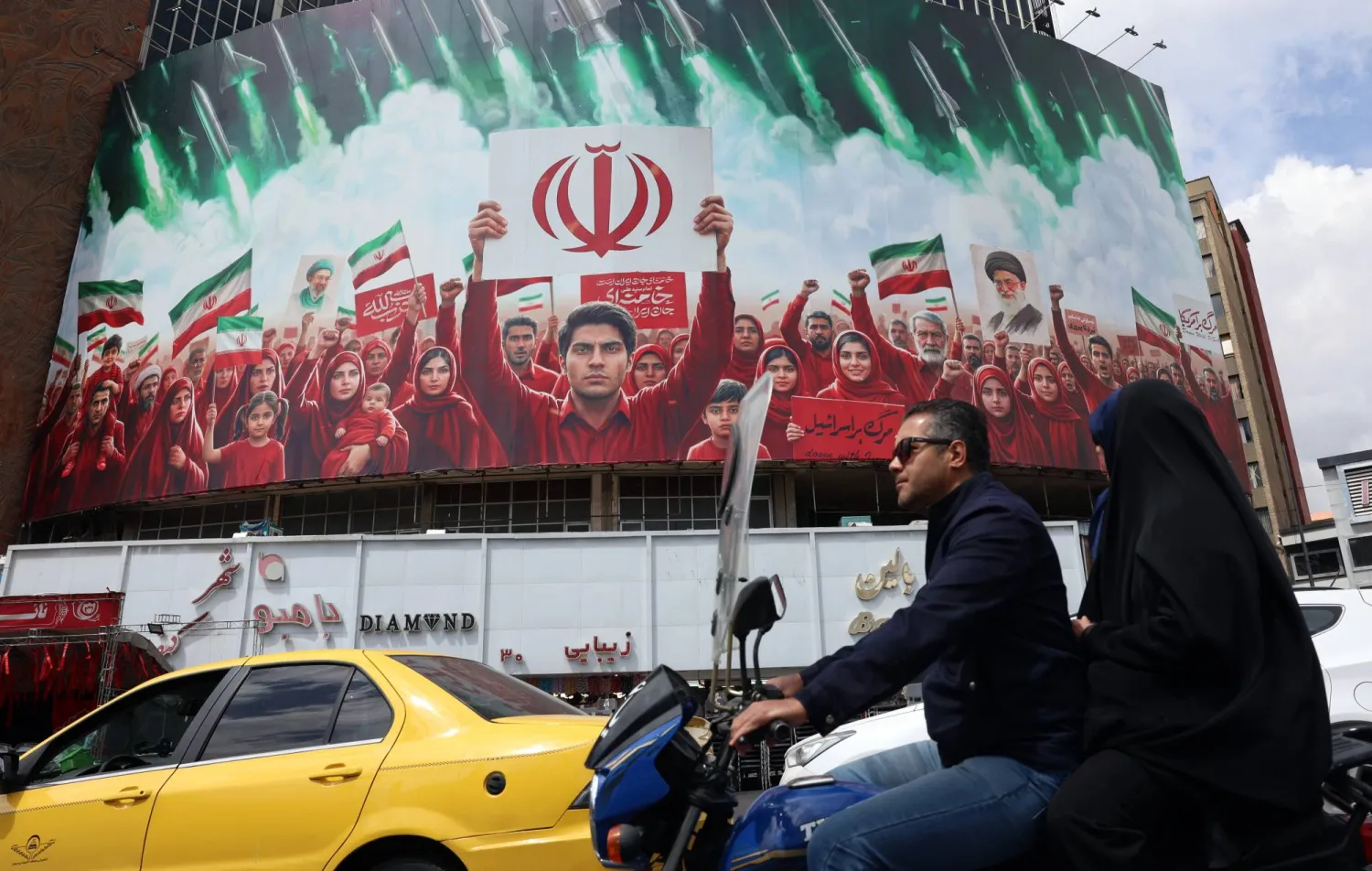 إيرانيون يمرون بسياراتهم أمام لوحة إعلانات سياسية تعرض أعلام إيران في إحدى ساحات العاصمة طهران 6 أبريل 2026 (إ.ب.أ)
