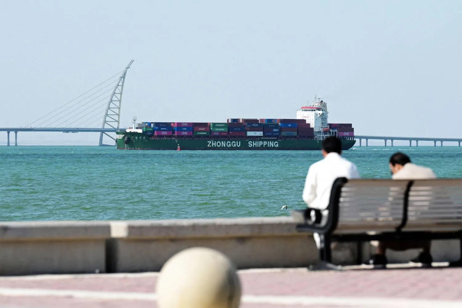 People sit on a bench on the Sheikh Jaber al-Ahmad al-Sabah Causeway as a Zhonggu Shipping container ship sails past on its way towards the port of Shuwaikh in Kuwait City on February 28, 2026. (Photo by YASSER AL-ZAYYAT / AFP)