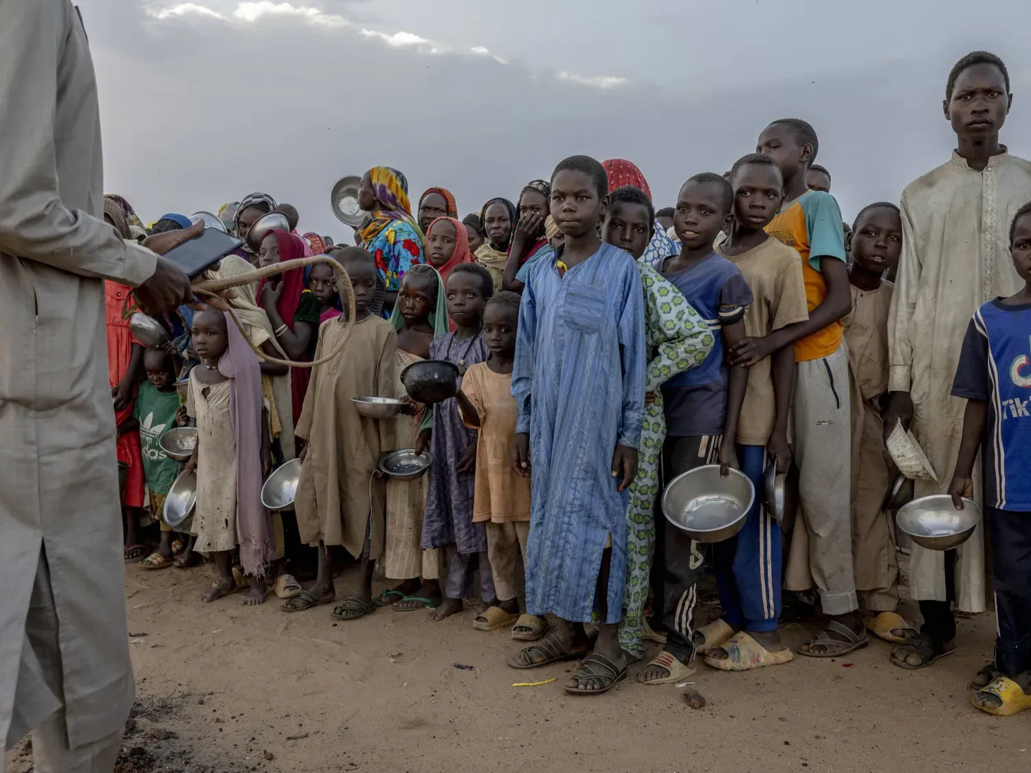 Sudan-Çad sınırında yer alan Adré (Adri) Mülteci Kampı’nda bir grup Sudanlı mülteci çocuk yemek almak için itişip kakışırken, elinde kırbaç tutan bir adam onları kontrol altına almaya çalışıyor (NYT)