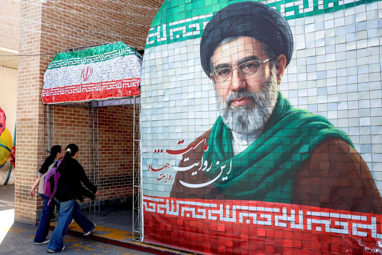 Women walk past a banner depicting Iran's current supreme leader Mojtaba Khamenei along a street Tehran on May 6, 2026. (Photo by AFP) 