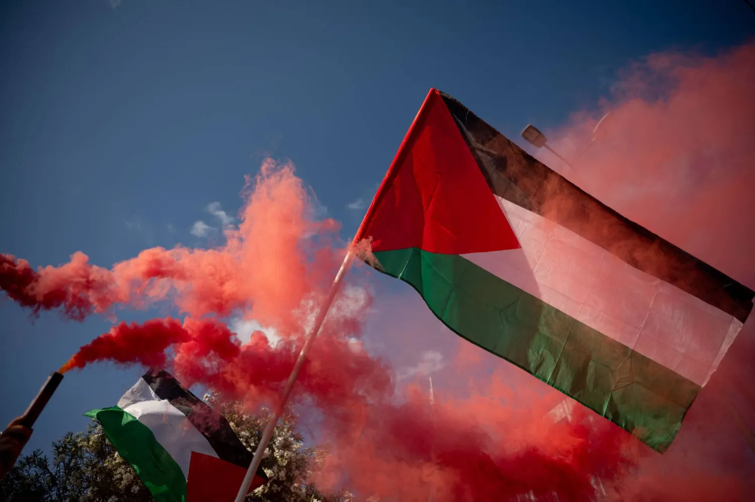 A Palestinian flag is pictured against a background of smoke grenades during a gathering against the interception of the Global Sumud Flotilla by the Israeli Navy off the coast of Cyprus outside the Ministry of Foreign Affairs in Athens on May 18, 2026.  (Photo by Aggelos NAKKAS / AFP)