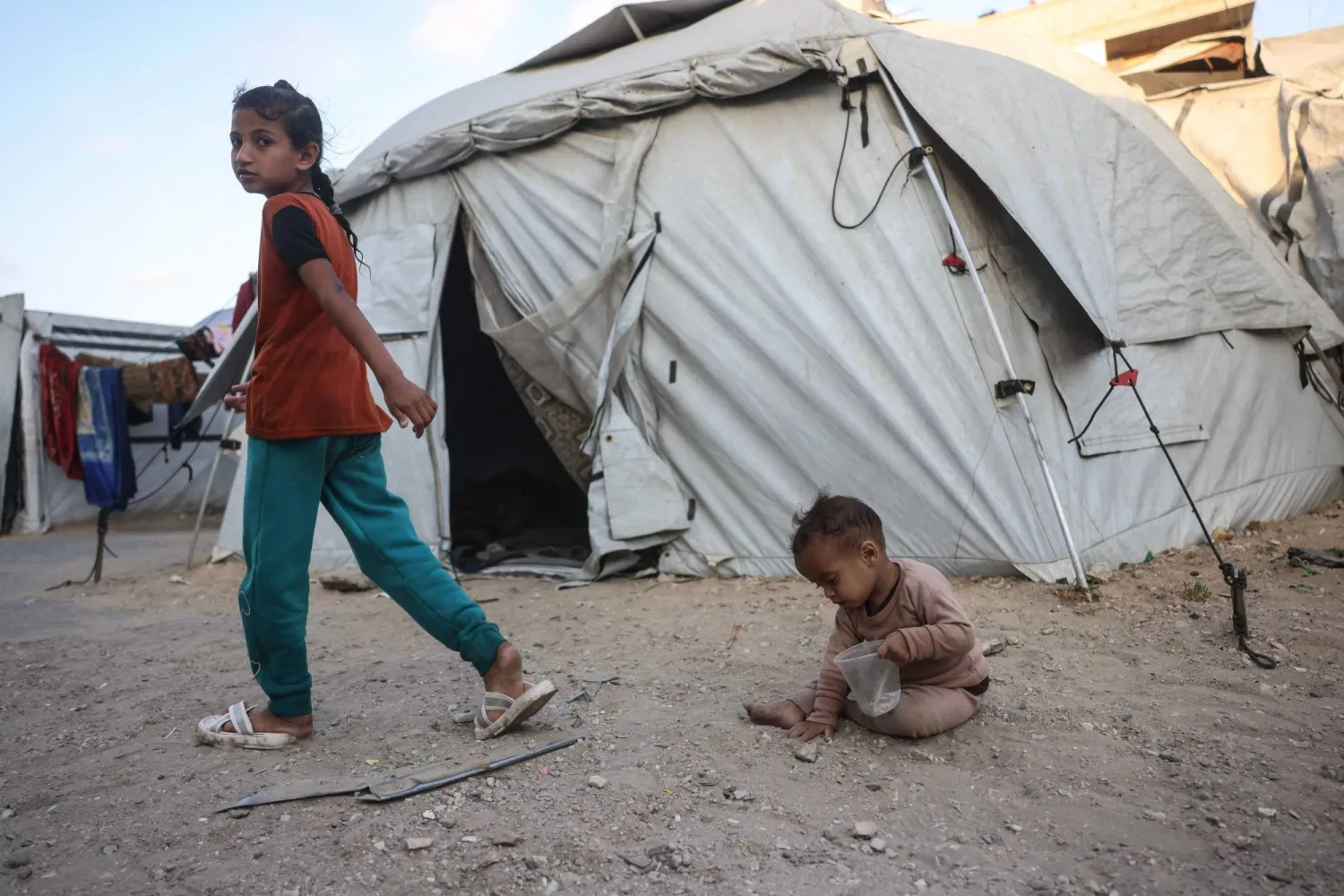A Palestinian girl walks past a toddler playing among makeshift shelters in Khan Yunis in the southern Gaza Strip on May 21, 2026. Since October 10, a fragile US-sponsored truce in Gaza has largely halted the fighting between Israeli forces and Hamas, but both sides have alleged frequent violations. (Photo by Bashar Taleb / AFP)