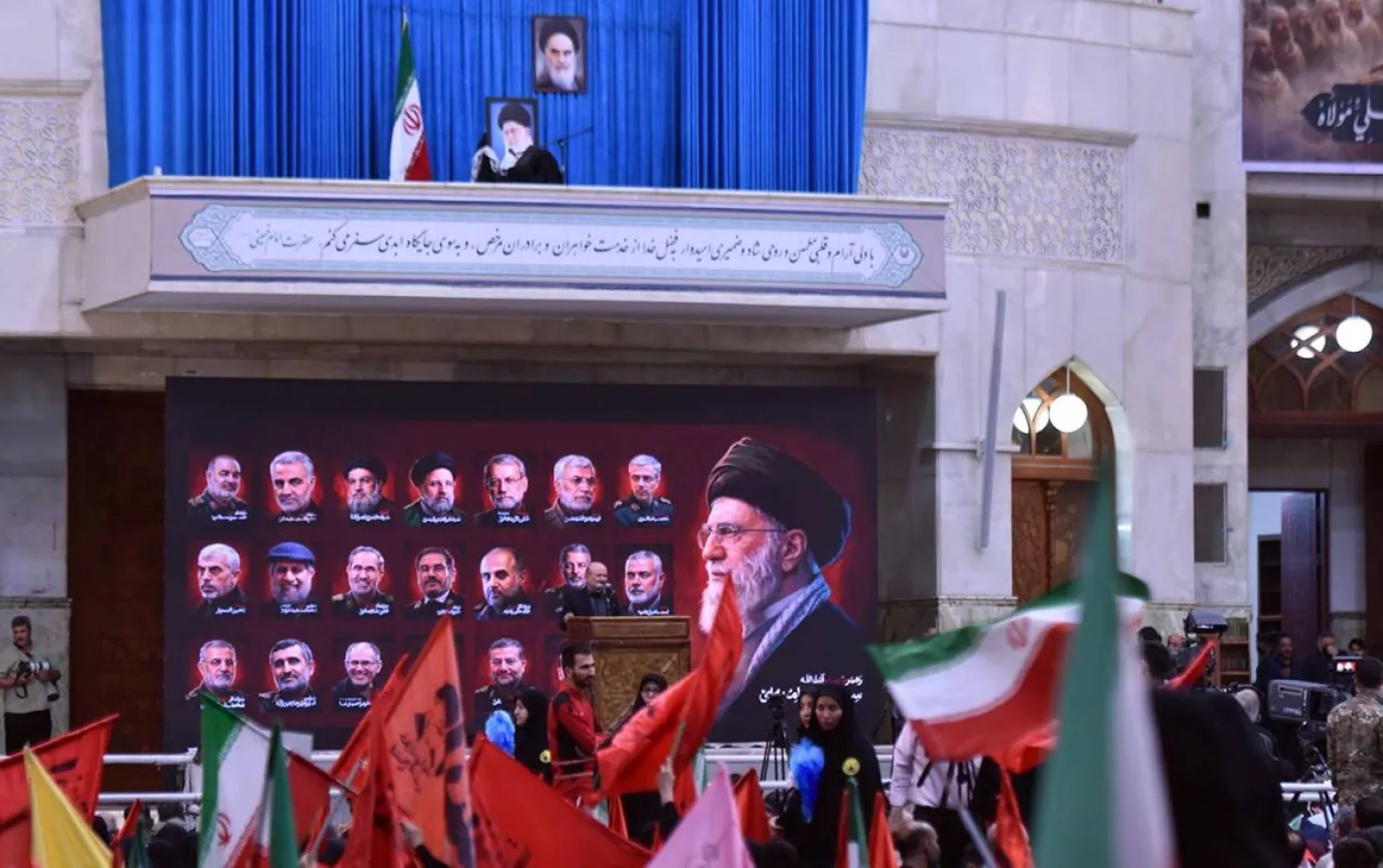 كرسي فارغ تعلوه صورة المرشد السابق علي خامنئي في موقع مراسم ذكرى وفاة الخميني جنوب طهران، وأدناها لوحة تضم صور قادة ومسؤولين إيرانيين قُتلوا خلال الحرب، بينما رُفعت أعلام الجمهورية الإسلامية ورايات حمراء بين الحشود المشاركة في المراسم (جماران)
