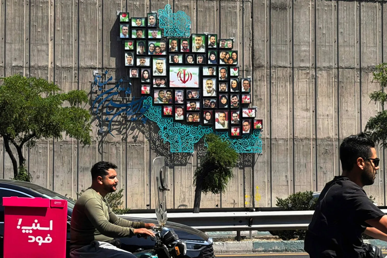 Men ride their mopeds past a map of Iran with the images of some of those killed in the Israeli-US war against the nation, erected on a wall along the highway in Tehran on June 17, 2026. (AFP)