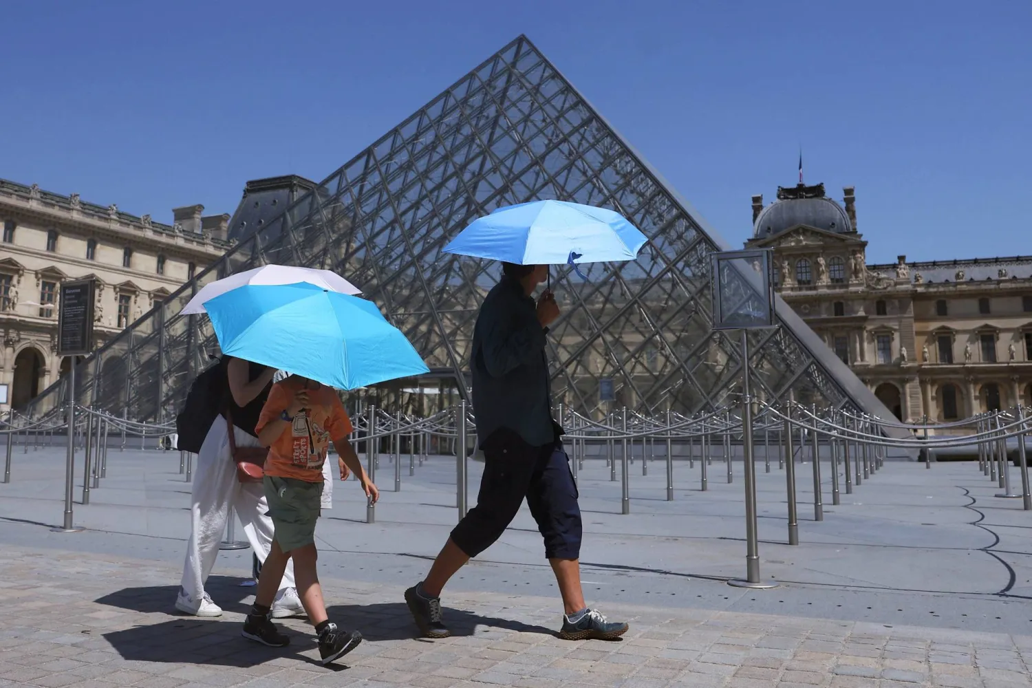 Tourists protecting themselves from the sun under umbrellas walk past the glass Pyramid of the Louvre museum as the museum closes early due to the heatwave as temperatures rise in Paris, France, June 24, 2026. REUTERS/Alice Sacco
