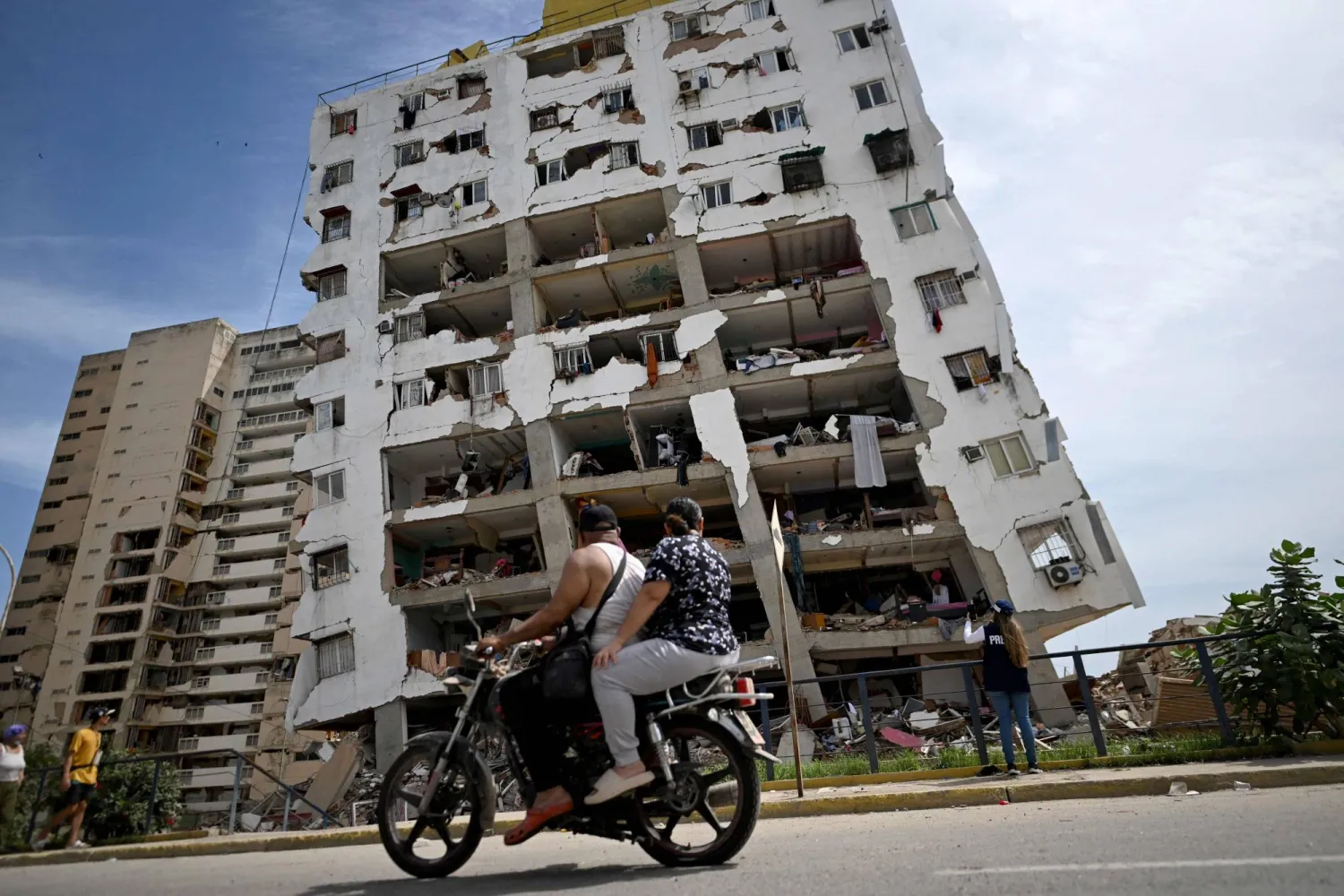 شخصان يقودان دراجة نارية أمام مبنى سكني متضرر بشدة وواجهته ممزقة إثر زلزال ضرب ولاية لا غوايرا على بعد نحو 30 كيلومتراً شمال غرب كراكاس (أ.ف.ب)