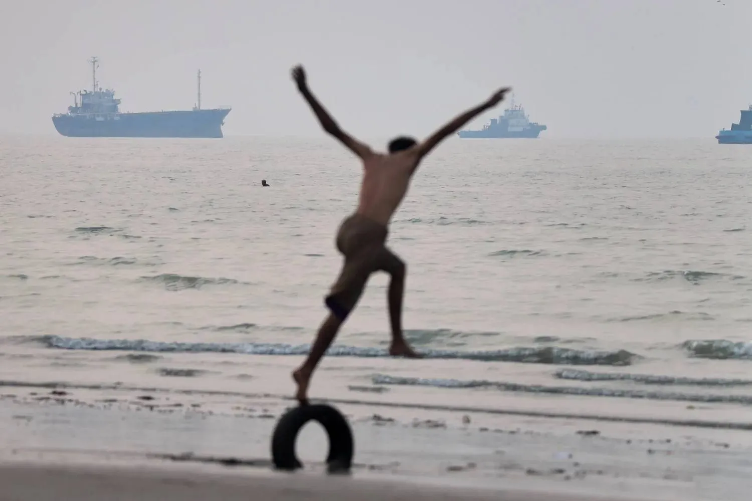 A boy balances on a tire at the water's edge as ships are seen in the Strait of Hormuz off Bandar Abbas, Iran, Tuesday, June 30, 2026 (AP) 