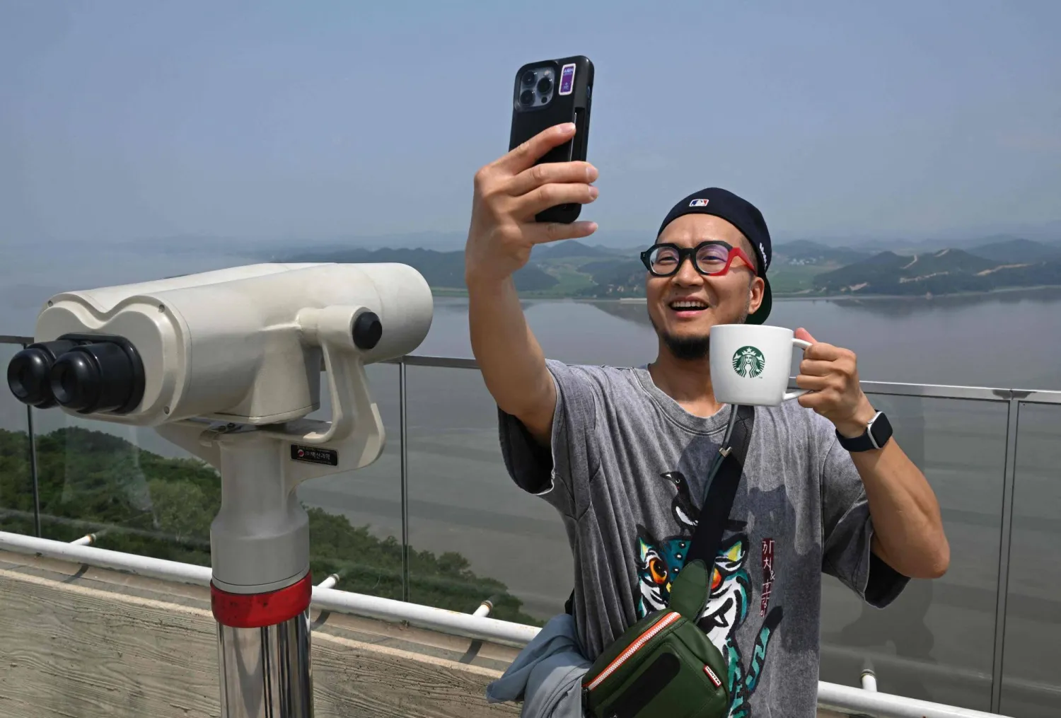 TOPSHOT - A visitor holding a Starbucks cup takes a selfie as he looks at the North Korean side from a South Korean observation deck at Aegibong Peace Ecopark in Gimpo on July 1, 2026. (Photo by Jung Yeon-je / AFP) 