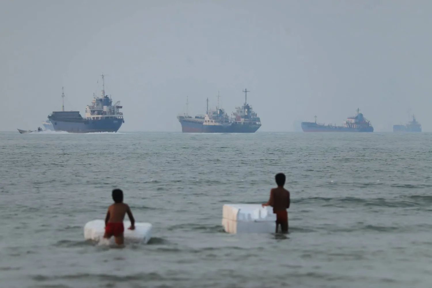 Two boys stand in shallow water with foam floats as cargo ships and other vessels are seen in the Strait of Hormuz off Bandar Abbas, Iran, Tuesday, June 30, 2026. (Amirhosein Khorgooi/ISNA via AP)