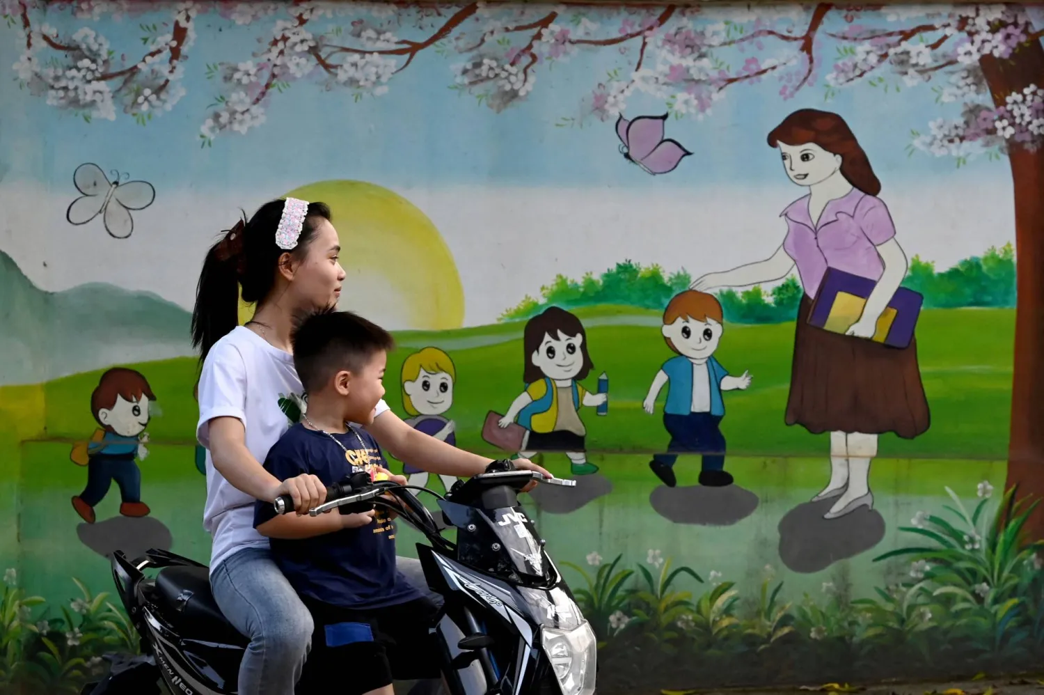 This photo taken on June 27, 2026, shows a young girl riding an electric motorbike with a child passing by a mural depicting a teacher welcoming children to school in Hanoi.(Photo by Nhac NGUYEN / AFP)
