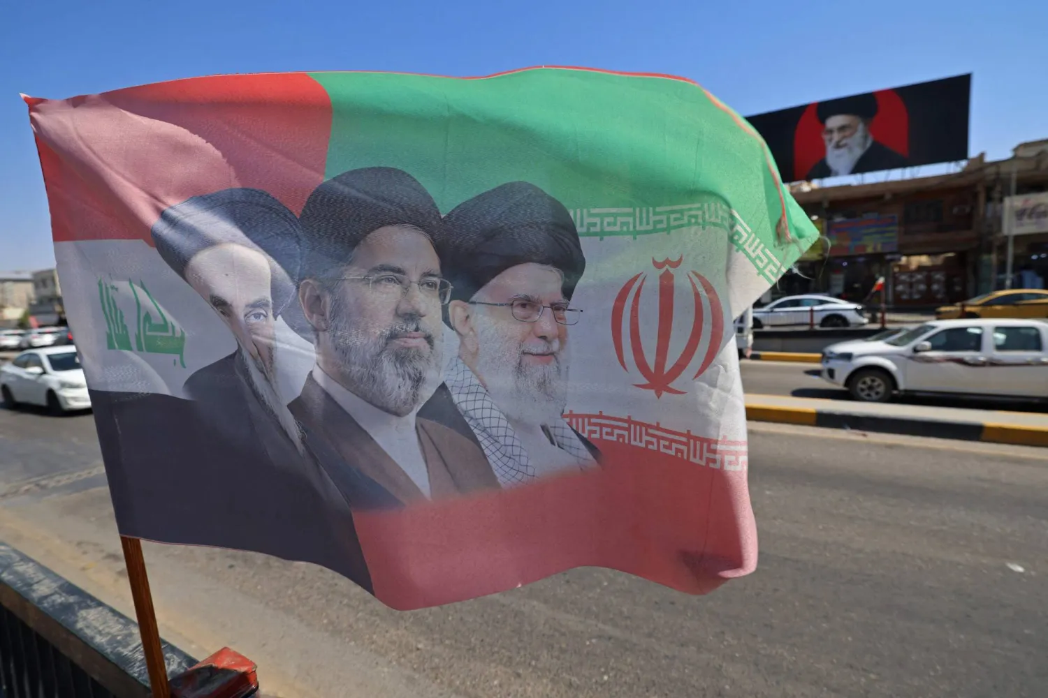 علم يحمل صور المرشد الإيراني الراحل علي خامنئي وابنه المرشد الحالي مجتبى خامنئي منصوباً على جانب أحد الشوارع في مدينة كربلاء بالعراق يوم 7 يوليو 2026 (أ.ف.ب)