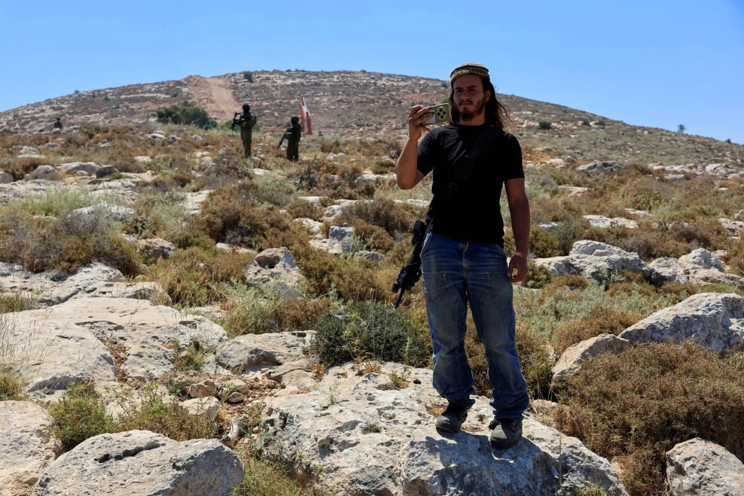 مستوطن إسرائيلي يلتقط صوراً بهاتفه في حين يقف جنود إسرائيليون حراساً خلال احتجاج فلسطيني ضد مستوطنة قرب الخليل في الضفة الغربية 9 يونيو 2026 (رويترز)
