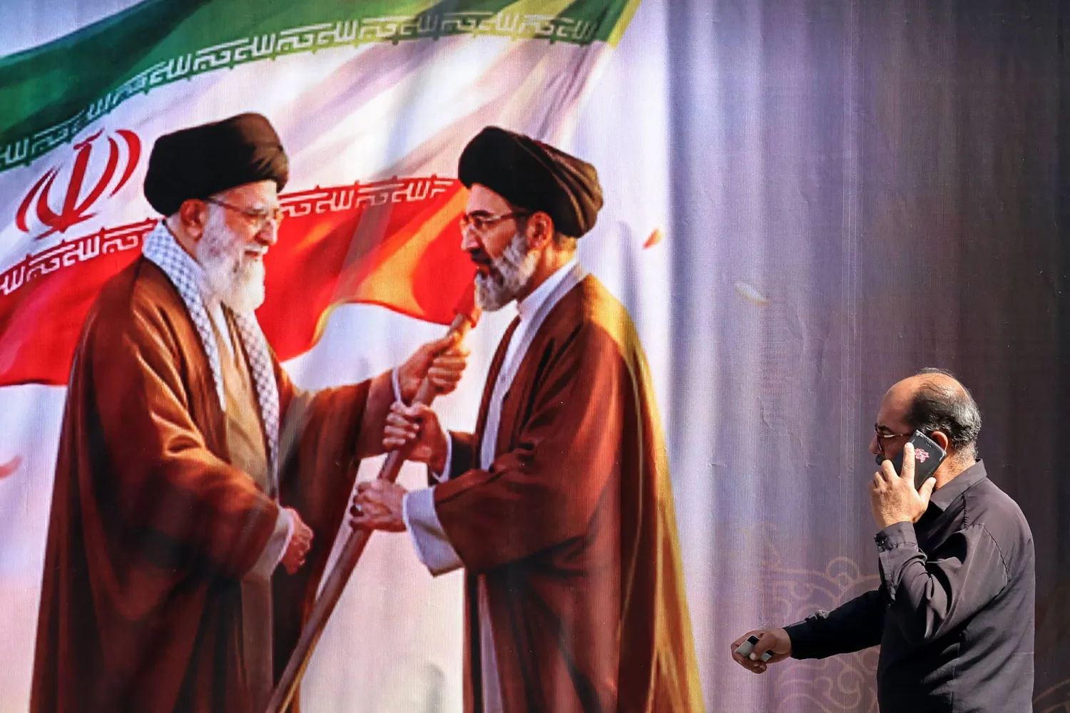 رجل يتحدث في الهاتف بينما يمر بجوار لوحة تظهر صورتي المرشدين السابق علي خامنئي (يسار) والحالي مجتبى خامنئي في طهران (أ.ف.ب)