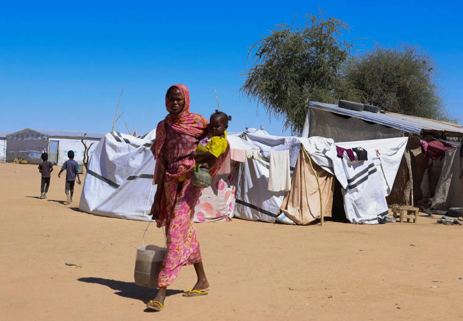 لاجئة سودانية من الفاشر تحمل طفلاً وماءً وخلفها خيام في مخيم «تين» للَّاجئين العابرين شرق تشاد (رويترز)