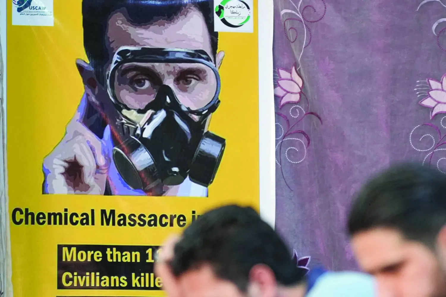 People sit across from a poster depicting Syria's President Bashar al-Assad in a gas mask during an event in the opposition-held northern city of Afrin, on August 20, 2023, marking the 10-year anniversary of chemical attacks that killed over 1,400 people in Ghouta, near the capital. (AFP)