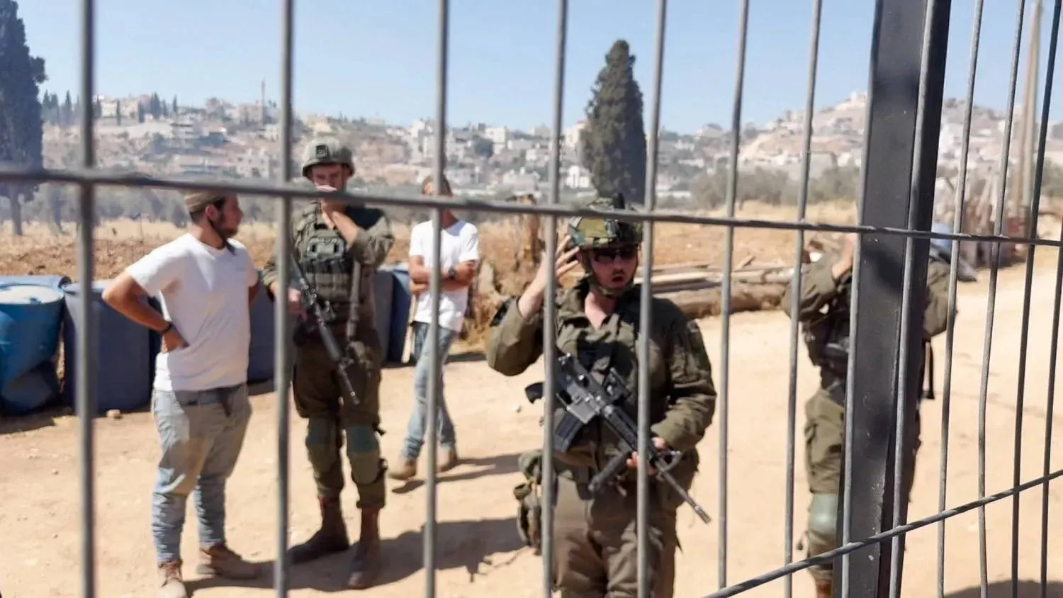 People wearing military uniform and Israeli settlers stand behind a Palestinian's home fence, during an Israeli settlers' attack on a Palestinian property in the village of Al-Mughayyir, near Ramallah, in the Israeli-occupied West Bank, July 11, 2026 (Reuters) 